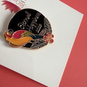 May include: A circular enamel pin with a black backdrop and gold text reading "Eternal Happiness 2026." The pin showcases a vibrant bird design, a red flower, and green leaves. The pin is presented on a white card.