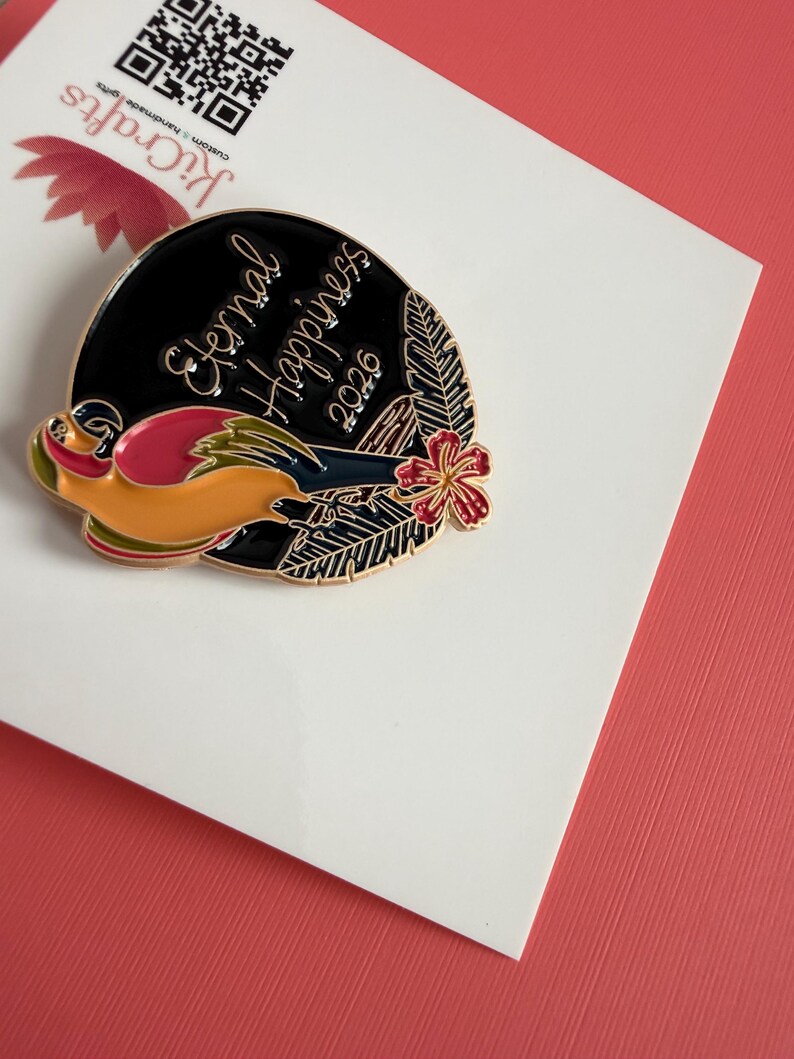 May include: A circular enamel pin with a black backdrop and gold text reading "Eternal Happiness 2026." The pin showcases a vibrant bird design, a red flower, and green leaves. The pin is presented on a white card.