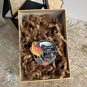 May include: A round enamel pin with a black border, featuring a colorful bird and the words "Eternal Happiness 2026." The pin is set in a cardboard box filled with brown packing material.