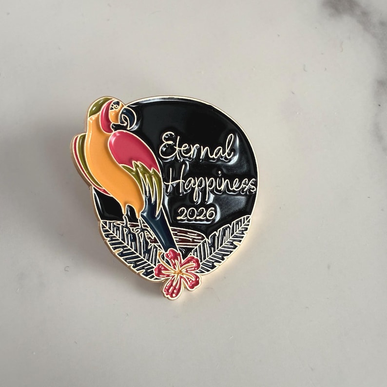 May include: A parrot-themed enamel pin with the words "Eternal Happiness 2026". The pin features a parrot in shades of orange, pink, and green, set against a black backdrop with a gold border and a red flower.