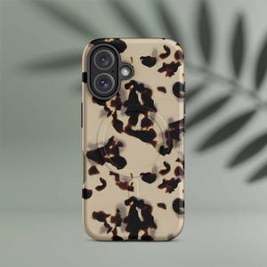Puede incluir: Una funda de teléfono con un diseño de carey en tonos beige y marrón oscuro. La funda tiene un accesorio magnético circular y un recorte para la cámara. La funda está diseñada para proteger el teléfono.