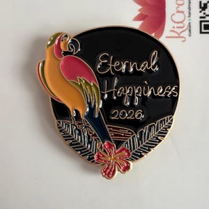 May include: A circular enamel pin with a parrot design. The parrot is yellow, pink, and blue, set against a black background with a red flower. The pin features the words "Eternal Happiness 2026" in gold script.