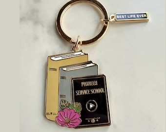 May include: A gold-toned metal keychain featuring a stack of three books in yellow, gray, and black. The black book is labeled PIONEER SERVICE SCHOOL with a play icon. A small rectangular tag attached to the ring reads BEST LIFE EVER in gold text.