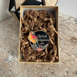 May include: A circular enamel pin featuring a parrot design in vibrant colours, with the text "Eternal Happiness 2026". The pin rests in a box with brown paper filling. A geometric wooden structure is in the background.