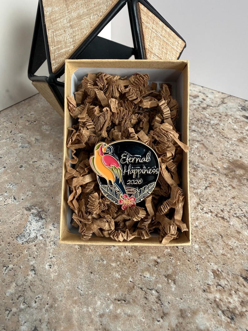 May include: A circular enamel pin featuring a parrot design in vibrant colours, with the text "Eternal Happiness 2026". The pin rests in a box with brown paper filling. A geometric wooden structure is in the background.