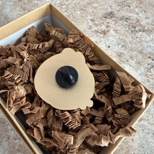 May include: A tan enamel pin with a black pin back, displayed in a small, square cardboard box filled with brown paper shreds. The pin has a rounded shape.