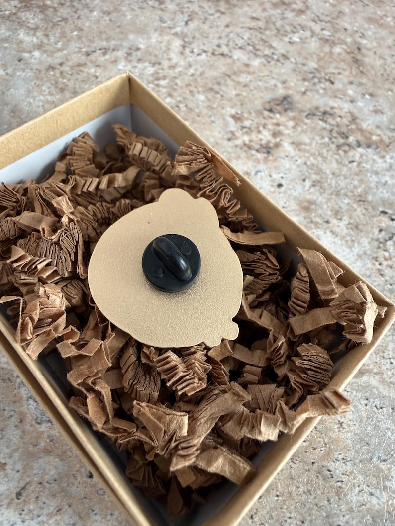 May include: A tan enamel pin with a black pin back, displayed in a small, square cardboard box filled with brown paper shreds. The pin has a rounded shape.