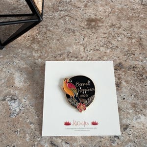 May include: A black and gold enamel pin featuring a parrot design and the words "Eternal Happiness 2020." The pin is on a white card with the KiCrafts logo and website address. The parrot is orange, yellow, and red.
