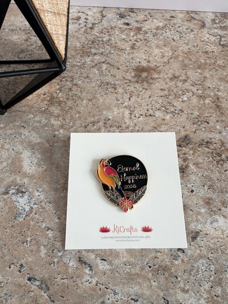 May include: A black and gold enamel pin featuring a parrot design and the words "Eternal Happiness 2020." The pin is on a white card with the KiCrafts logo and website address. The parrot is orange, yellow, and red.