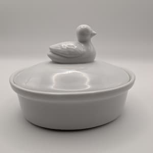 May include: A white ceramic lidded dish featuring a sculpted duck figurine as the handle on the lid. The container is round with smooth, glossy surfaces and a simple, clean design, showcasing a uniform white finish throughout the entire piece.