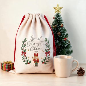 May include: A cream-coloured drawstring gift bag with red cords, featuring a printed nutcracker, holly branches, and the text Special Delivery For Chloe 2026. The bag is displayed alongside a small Christmas tree, a gift box, and a mug.