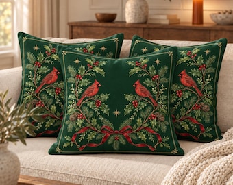 May include: A set of three square scatter cushions in a deep forest green colour. Each cushion features a printed design of two red cardinal birds perched on holly branches with red berries, pine cones, and a red ribbon bow at the bottom.