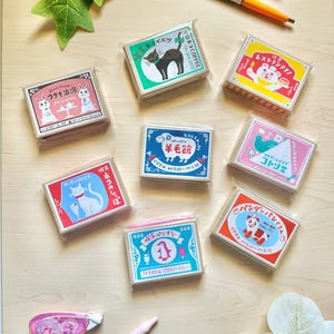 May include: Eight rectangular memo pads in clear plastic packaging, each featuring unique Japanese graphic illustrations of animals like cats, rabbits, bears, sheep, birds, and pandas, arranged on a light wood surface with a pen and tape dispenser.