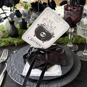 Può includere: Una carta rettangolare bianca con disegni a linee nere raffiguranti un calderone con un teschio, vapore e il nome Stephanie stampato in un carattere decorativo. La carta è legata con un nastro nero a un fascio di tessuto su un piatto nero.