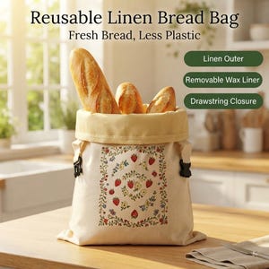 May include: A cream-colored reusable linen bread bag featuring a rectangular floral and strawberry print. The bag has a drawstring closure, a removable wax liner, and contains three loaves of bread. Text reads: Reusable Linen Bread Bag, Fresh Bread, Less Plastic, Linen Outer, Removable Wax Liner, Drawstring Closure.
