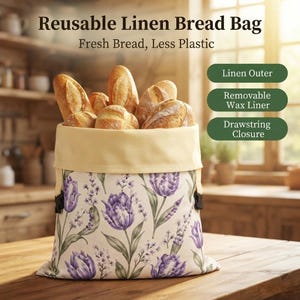 May include: A reusable linen bread bag with a cream-colored top and a floral pattern of purple tulips and lavender on a white background. The bag is filled with various crusty bread loaves and features text: Reusable Linen Bread Bag, Fresh Bread, Less Plastic, Linen Outer, Removable Wax Liner, Drawstring Closure.