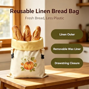 May include: A reusable linen bread bag with a cream-colored exterior featuring a pumpkin illustration. The bag contains three baguettes and has a yellow interior lining, black drawstring closures, and text reading Reusable Linen Bread Bag, Fresh Bread, Less Plastic, Linen Outer, Removable Wax Liner, and Drawstring Closure.