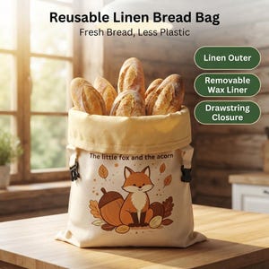 May include: A reusable linen bread bag with a drawstring closure and a removable wax liner. The bag is cream-colored and features a graphic of a fox and an acorn with the text "The little fox and the acorn." It is filled with several loaves of bread.