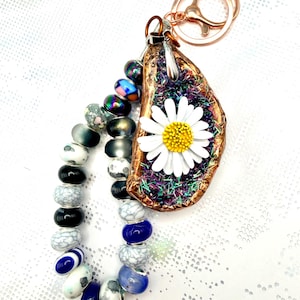 May include: A keyring featuring a rose gold ring attached to a decorative oyster shell with a white daisy flower centre. The shell interior is filled with purple glitter and iridescent fibres. A strand of assorted beads in blue, white, black, and metallic tones hangs alongside the shell.