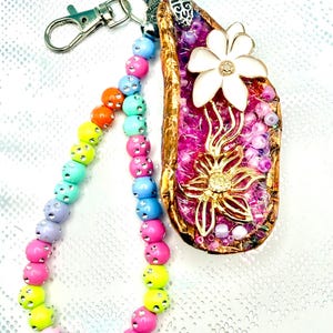 May include: A keyring featuring a gold-rimmed, teardrop-shaped pendant filled with bright pink glitter, a white enamel flower, and a gold-tone metal flower. A wrist strap made of multi-coloured, rhinestone-studded beads is attached to a silver metal clasp.