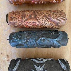 Pode incluir: Quatro rolos de pintura cilíndricos com padrões dispostos verticalmente. O rolo superior é laranja brilhante com motivos florais. O segundo é bege mate com desenhos botânicos intrincados. O terceiro é cinzento escuro com padrões de folhas. O inferior é cinzento escuro com uma estampa floral branca.