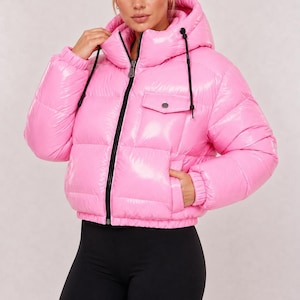 May include: A bright pink, glossy puffer jacket featuring a high collar, a hood with black drawstrings, a front zipper closure, and a single chest pocket with a flap and button. The jacket has long sleeves with elastic cuffs and a cropped hemline.
