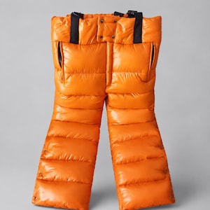 Può includere: Pantaloni imbottiti color arancione brillante con design a vita alta e bretelle nere regolabili. I pantaloni presentano una struttura trapuntata e isolante con finitura lucida, due tasche laterali e chiusure con bottoni a pressione.