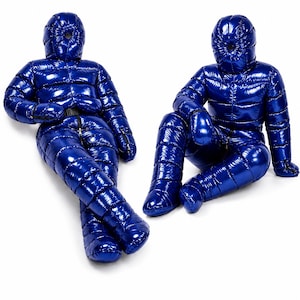Puede incluir: Dos trajes de cuerpo completo acolchados en color azul real brillante con capuchas y manoplas integradas. Los trajes presentan una construcción de deflectores horizontales, una cremallera central frontal y un cinturón negro. Uno está tumbado y otro sentado.