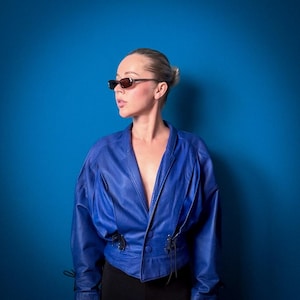 May include: A vibrant blue leather jacket featuring a deep V-neckline, two large matching buttons at the centre, and pleated detailing on the front panels. The jacket includes thin black lace-up accents at the waist and dark-tinted rectangular sunglasses.