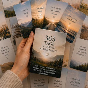 Könnte beinhalten: Ein Set aus Affirmationskarten mit Naturfotografien und deutschem Text. Die Box trägt den Titel 365 TAGE ZU DEINEM NEUEN LEBEN vor einer Bergkulisse. Jede Karte enthält einen individuellen deutschen Satz und ein kleines Herzsymbol als Verzierung.