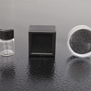May include: A small clear glass vial with a black screw-top lid, a square black display box with a foam insert, and a round clear plastic container filled with a fine dark gray granular substance, arranged in a horizontal row.