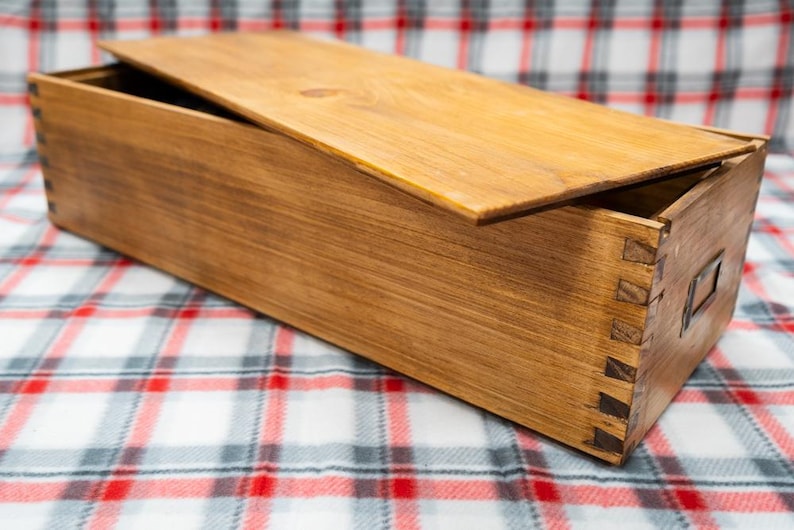 May include: A rectangular wooden storage box with a natural brown finish and visible finger joint construction at the corners. The box features a flat, removable wooden lid and a small metal label holder attached to one of the short side panels.