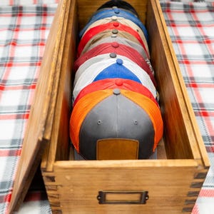 May include: A rectangular wooden storage box with finger-jointed corners and a metal label holder on the front, containing a vertical stack of multiple baseball caps in various colors including grey, orange, red, blue, and white with mesh panels.