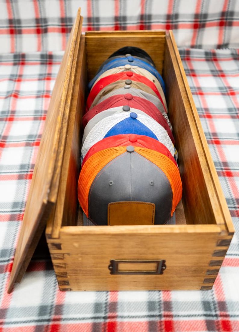May include: A rectangular wooden storage box with finger-jointed corners and a metal label holder on the front, containing a vertical stack of multiple baseball caps in various colors including grey, orange, red, blue, and white with mesh panels.