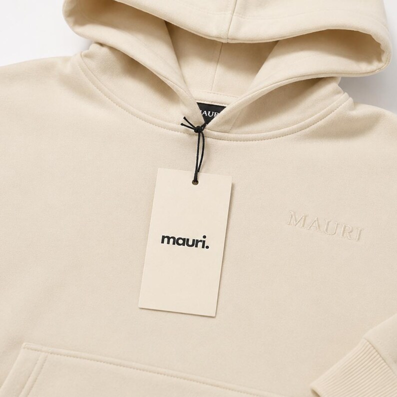 May include: A cream-colored hooded sweatshirt featuring a matching drawstring hood and a front kangaroo pocket. The word MAURI is embroidered on the chest, and a rectangular tag with the text mauri. is attached to the neckline with a black cord.