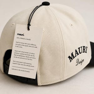 May include: A cream-colored baseball cap with a black button on top and a black snapback closure. The side features black embroidered text reading MAURI Design. A rectangular off-white tag is attached, printed with the text mauri. not a brand. a world.