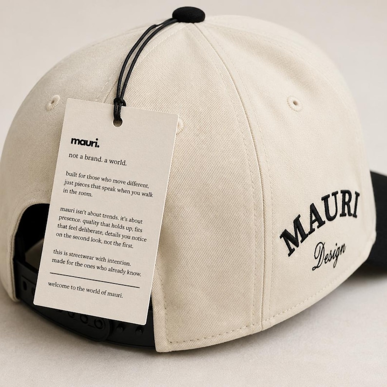 May include: A cream-colored baseball cap with a black button on top and a black snapback closure. The side features black embroidered text reading MAURI Design. A rectangular off-white tag is attached, printed with the text mauri. not a brand. a world.