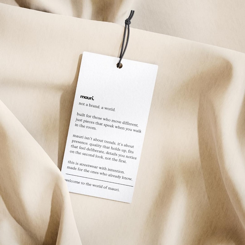 May include: A rectangular white paper hang tag with black text attached by a thin black string. The text reads: mauri. not a brand. a world. built for those who move different, just pieces that speak when you walk in the room. welcome to the world of mauri.