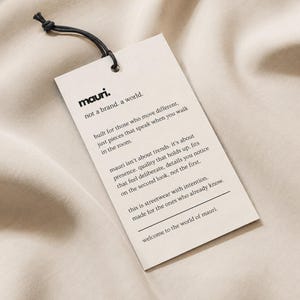 May include: A rectangular off-white paper hang tag with a black string loop. The tag features the black printed text: mauri. not a brand. a world. built for those who move different, just pieces that speak when you walk in the room.