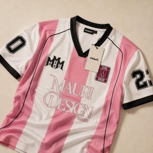 May include: A short-sleeved V-neck jersey with vertical pink and white stripes, black trim, and the text Mauri Design Est. 2025. The sleeves feature the numbers 0 and 23 in black. A white tag with the text mauri. is attached to the collar.