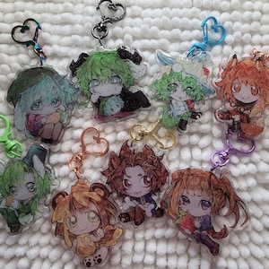 Puede incluir: Una colección de ocho llaveros de acrílico con coloridas ilustraciones de personajes estilo anime. Cada dije está sujeto a un broche de metal en forma de corazón con varios acabados, incluyendo iridiscente, negro, azul, oro rosa, dorado y morado.