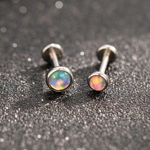May include: Two silver-toned labret studs featuring round, iridescent opal cabochons. Each stud has a smooth metal post and a flat back disc. The opals display a play of color with flashes of blue, green, and pink against a metallic setting.