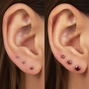 May include: A set of three round, red gemstone stud earrings arranged in a vertical line on an earlobe. The earrings feature a polished metal bezel setting, with the size of the stones increasing progressively from the top to the bottom position.