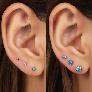 May include: A set of three stud earrings featuring round, light blue gemstones set in silver-toned metal. The earrings are arranged in a row along the earlobe, with each stud progressively increasing in size from the smallest to the largest.
