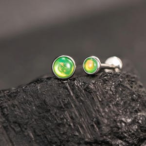 May include: A pair of circular stud earrings featuring smooth, polished metal settings and vibrant, colour-shifting cabochon stones that display gradients of bright green and yellow. The earrings have a metallic post back and a rounded, dome-like profile.
