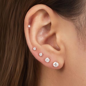 May include: A set of five round, clear, brilliant-cut stud earrings arranged in a vertical line along the earlobe and cartilage. Each earring features a circular gemstone set in a gold-toned metal prong setting, graduating in size from smallest to largest.