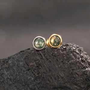 May include: Two round stud earrings featuring moss agate cabochons with green and translucent inclusions. One earring is set in a polished silver-tone metal bezel, while the other is set in a polished gold-tone metal bezel, both displaying a smooth finish.