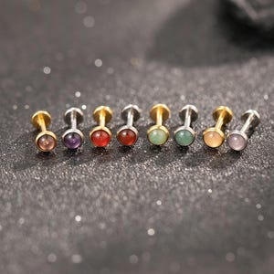 Könnte beinhalten: Eine Reihe von acht Ohrsteckern mit Metallstiften und runden, cabochonartigen Edelsteinen in verschiedenen Farben wie Violett, Rot, Orange, Grün und Hellrosa, die horizontal auf einer dunklen, strukturierten Oberfläche angeordnet sind.