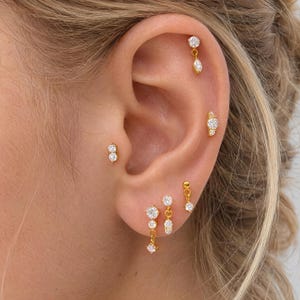 May include: A collection of gold-tone earrings featuring clear, round, and marquise-cut stones. The set includes multiple stud and dangling designs worn in various ear piercings, showcasing a metallic finish and sparkling white accents.