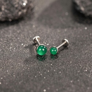 May include: Two silver-toned labret studs featuring smooth, round, vibrant green gemstone beads as the front decorative element. Each piece consists of a straight metal post with a flat back and a polished green sphere securely attached to the front.
